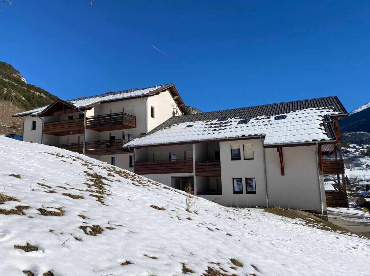Ein modernes Gebäude mit mehreren Balkonen und einem mit Schnee bedeckten Dach, gelegen an einem Hang unter blauem Himmel.