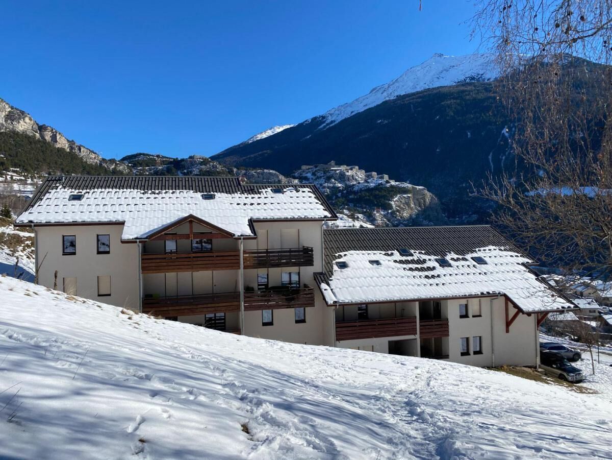A multi-story chalet with a sloped roof, located on a snowy hillside with a mountain backdrop.