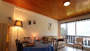 Uma sala de jantar com uma mesa redonda, cadeiras e toalha de mesa azul. Há uma luminária à esquerda, um jarro e copos sobre a mesa. O ambiente possui paredes e teto de madeira, com uma janela e varanda visíveis.