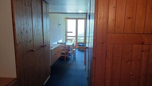 A narrow hallway with wooden paneling, leading to a room with a table and chairs, and a large window with a view of greenery.