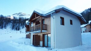 Un chalet à deux étages doté d’un balcon en bois, avec une piste de ski en arrière-plan et un paysage enneigé.