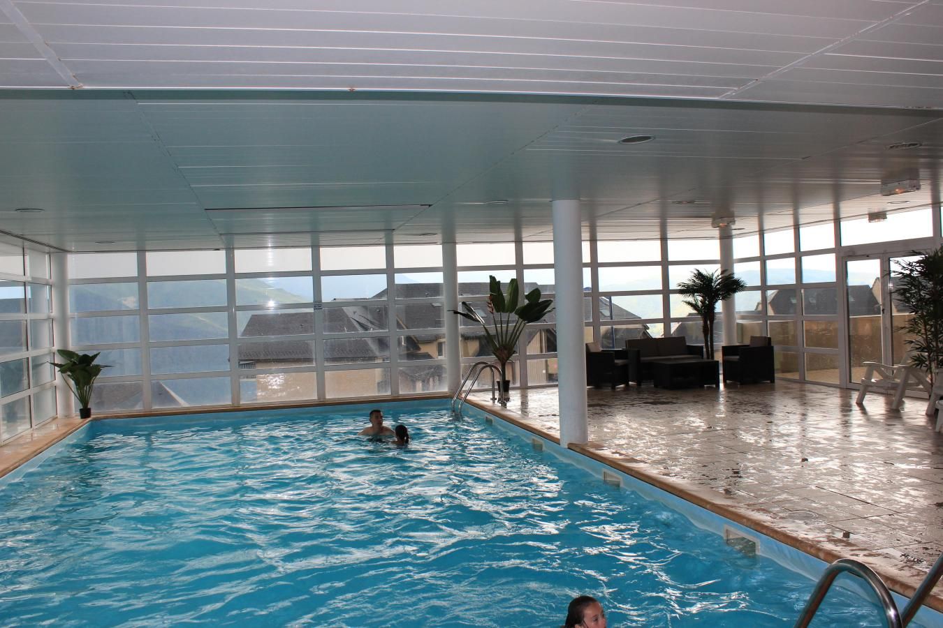 An indoor swimming pool with clear glass walls, a view of the sea, and a seating area with chairs and tables.