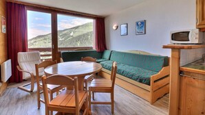 A cozy room with a wooden dining table, chairs, a sofa, and a view of a mountainous landscape through large windows. - 10m from ski slopes, 5th floor, balcony, 33m², Méribel-Mottaret (Méribel - Les Allues)