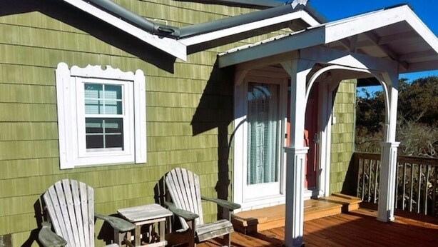 A wooden deck with two Adirondack chairs and a small table, a covered porch area, and a window with white trim. - The Lucky Buc-Pet friendly cottage perfect for a couple or small family. (Ocracoke)