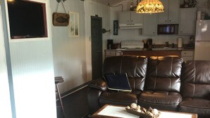 A living room with a brown leather sofa, a wooden coffee table, and a woven ottoman. A kitchen is visible in the background with white cabinets and a microwave. - The Lucky Buc-Pet friendly cottage perfect for a couple or small family. (Ocracoke)