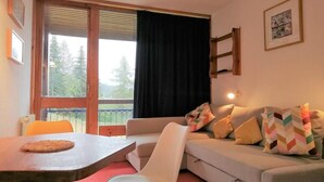 Una sala de estar con sofá, una mesa de comedor y una silla. Se ve una ventana con vistas a árboles.