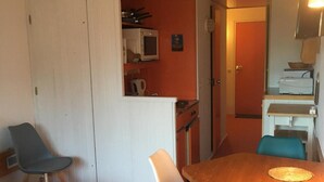 Un espacio habitable compacto con comedor que incluye una mesa de madera y cuatro sillas, una kitchenette con microondas, y una pequeña zona de estar con un sofá y dos sillas.