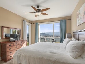 A bedroom with a bed, a wooden dresser, a ceiling fan, a TV, and a view of boats on the water through large windows.
