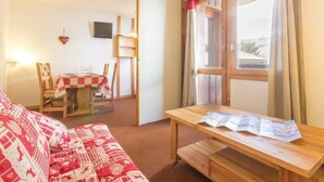 Una habitación acogedora con un sofá rojo estampado, una mesa de madera con folletos, una mesa de comedor de madera con mantel a cuadros y una ventana con cortinas.