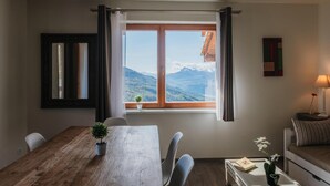 Een kamer met een houten eettafel, stoelen, een raam met uitzicht op een berg, een kleine plant op de vensterbank, een bank met een kussen en een lamp op een bijzettafeltje.