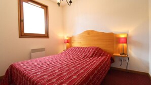 A bedroom with a wooden headboard, a red patterned bedspread, two bedside lamps, and a window with blinds.