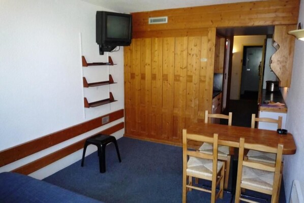 A room with a wooden wall, a television mounted above a shoe rack, a dining table with chairs, and a blue carpet.
