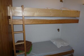 A bunk bed with a wooden ladder, a single bed below, and a small shelf with a green bag.