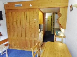 A room with a wooden dining table, chairs, a kitchen area with a stove, and a blue carpet.