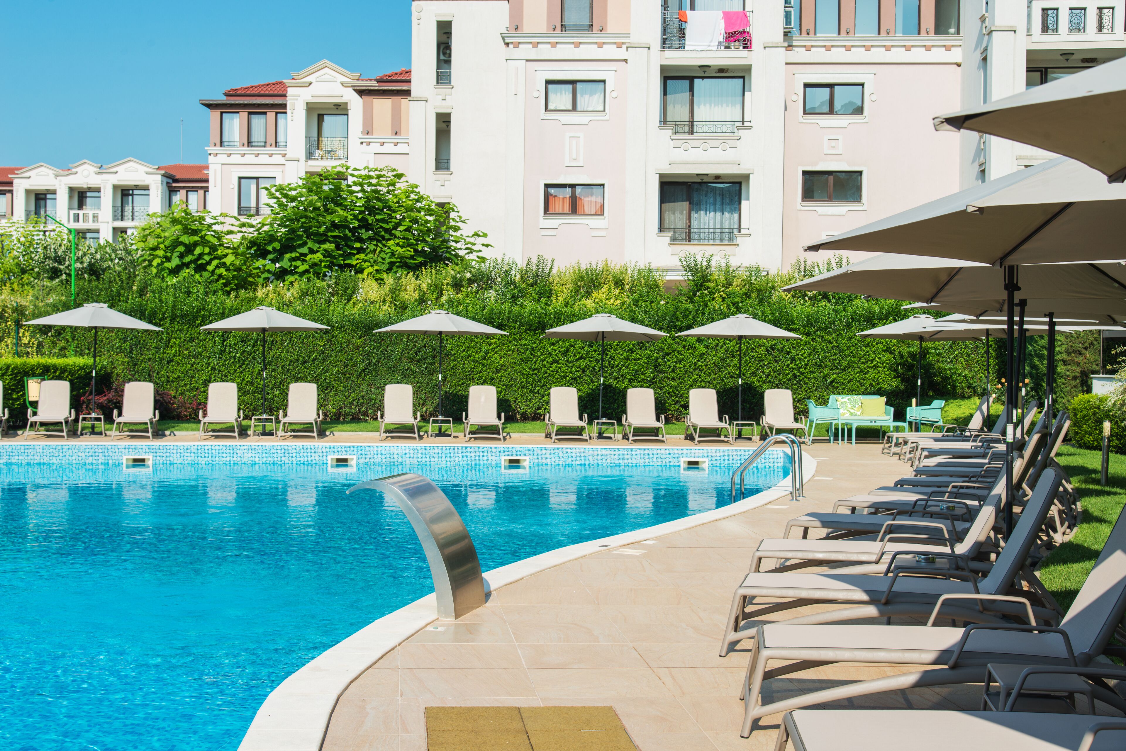 Piscina con sillones de descanso y sombrillas, rodeada de vegetación y un edificio con balcones.