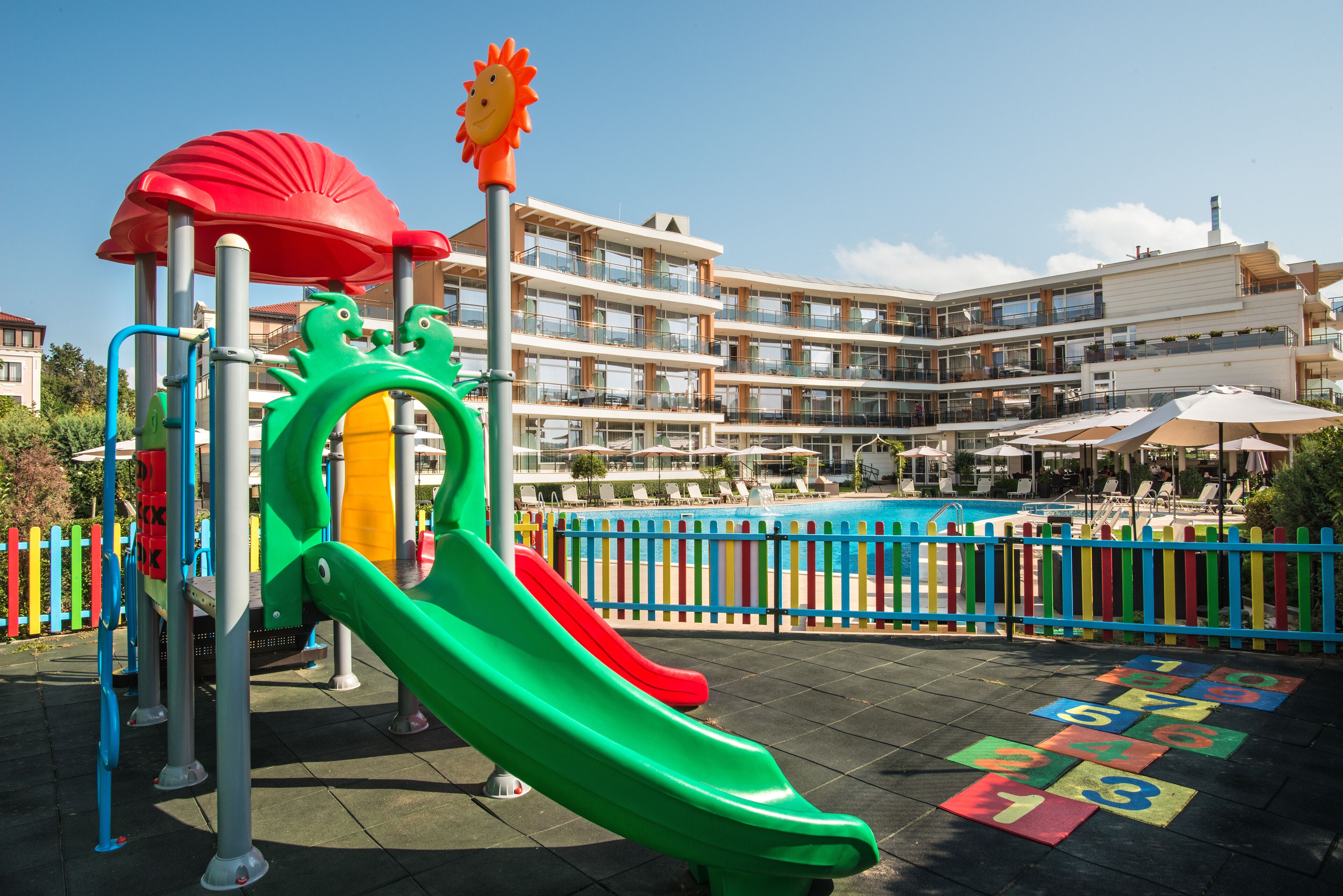 Ein Spielplatz mit Rutschen und Klettergerüsten, ein Poolbereich mit Liegestühlen sowie ein mehrstöckiges Gebäude mit Balkonen.