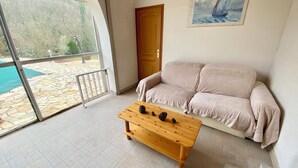 A living room with a beige sofa, a wooden coffee table with pinecones, a painting of a sailboat, and a view of a stone patio and pool through a glass door. - Vacation rental for 2–4 people, 1 bedroom, 1 shower room, heated outdoor pool, garden, trampoline (Ardèche)
