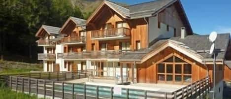 A multi-story wooden chalet with balconies, a swimming pool, and a mountainous backdrop.