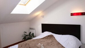 Une chambre avec un puits de lumière, un lit avec une literie blanche, un jouet en peluche marron, une plante en pot et un luminaire rouge.