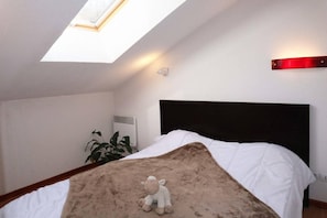 A bedroom with a skylight, a bed with white bedding, a red wall-mounted light, and a plant.