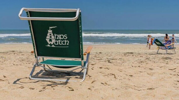Uma cadeira de praia com um logotipo no encosto, colocada numa praia arenosa com o oceano ao fundo.