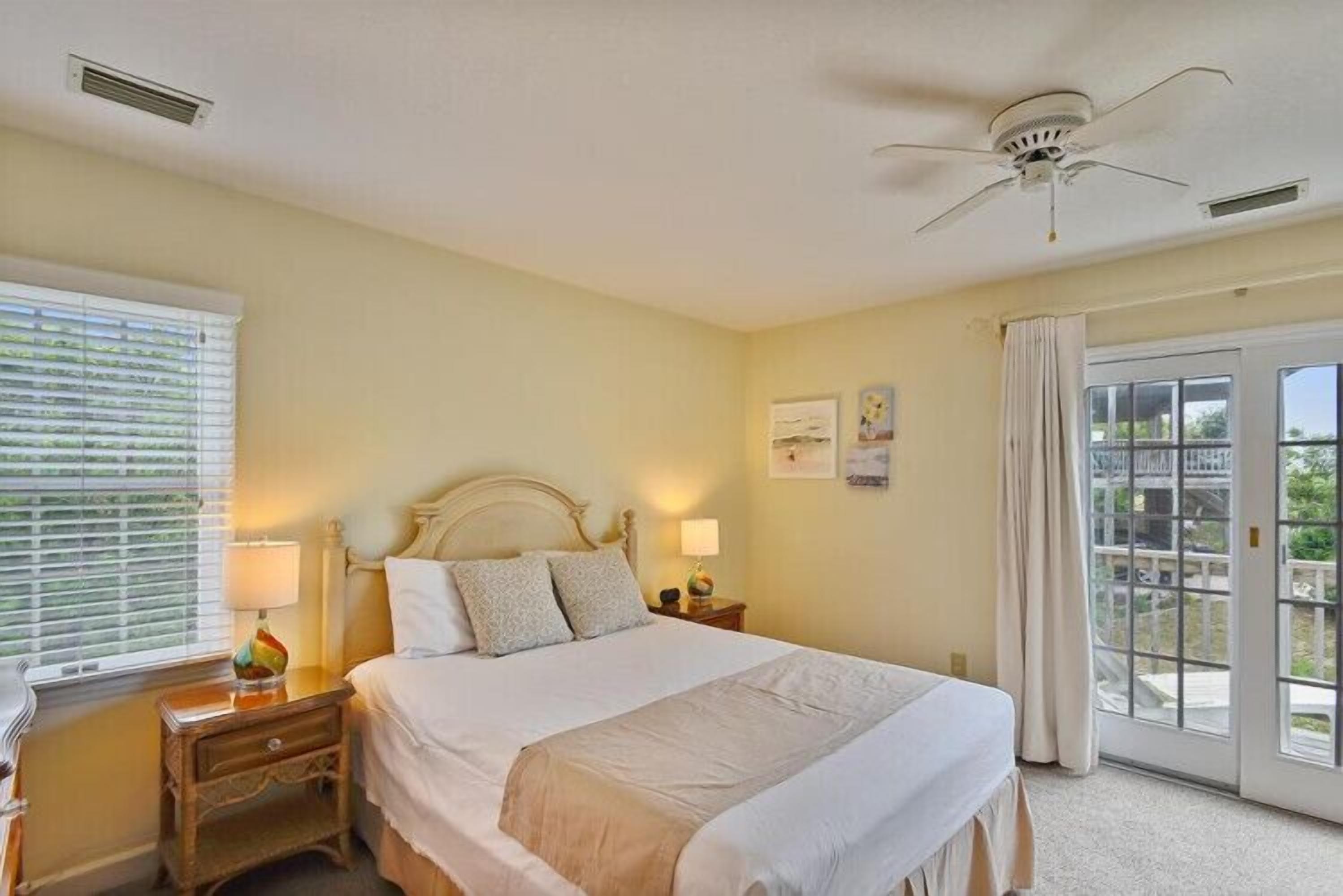 Une chambre avec un grand lit, un ventilateur de plafond, une table de chevet avec une lampe et une fenêtre avec des stores.