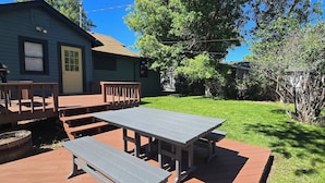 A patio area with a table and benches, a small deck, and a house in the background. - Cottonwood Cottage- Walk to historic Downtown Cody, private yard (Cody)