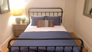 A bed with a metal frame, a wooden nightstand with a lamp, and a small plant. - Cottonwood Cottage- Walk to historic Downtown Cody, private yard (Cody)