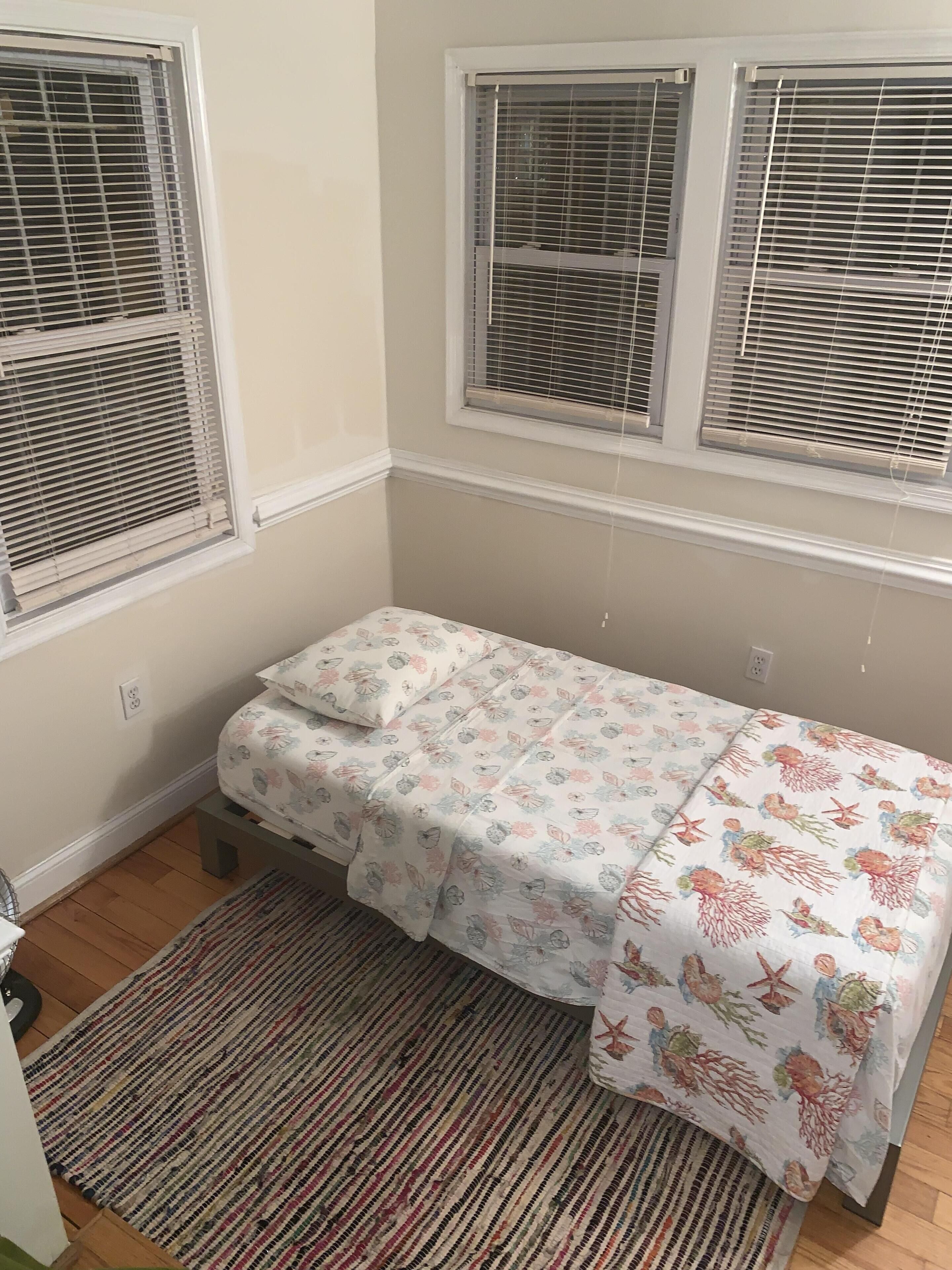 A small, single-bed room with a floral-patterned bedspread, a striped rug, and three windows with blinds.