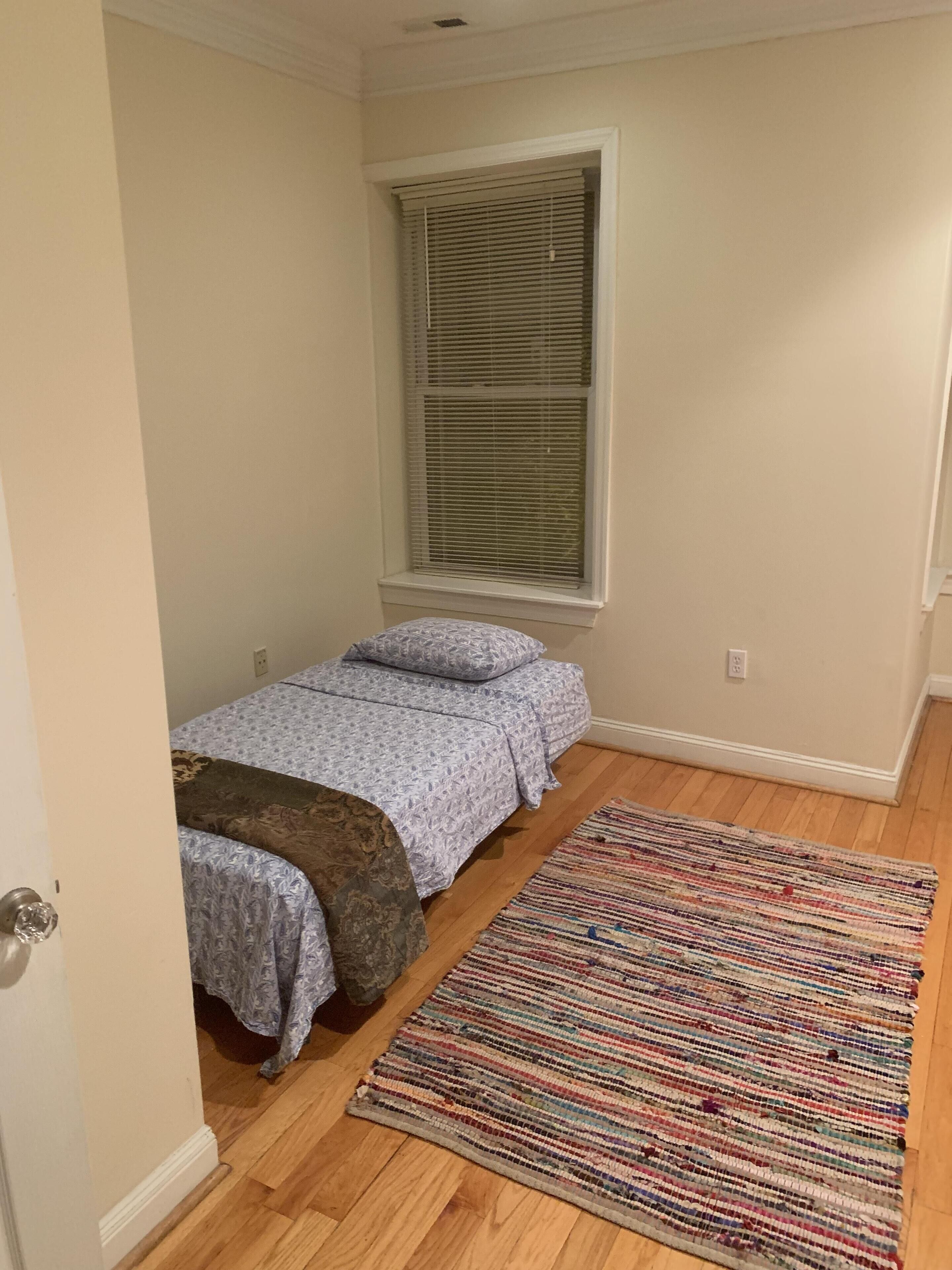 A small bedroom with a single bed, a window with blinds, a colorful rug, and wooden flooring.