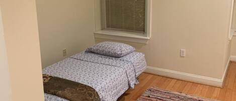 A small bedroom with a single bed, a window with blinds, a colorful rug, and wooden flooring.