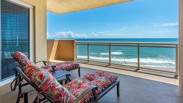 Un balcon avec deux fauteuils et une table, donnant sur une plage où l’on peut voir des vagues et des gens.