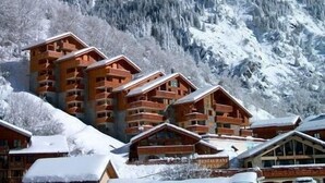 Un albergue de montaña con varias cabañas de madera, un letrero que indica un restaurante y una montaña nevada al fondo.