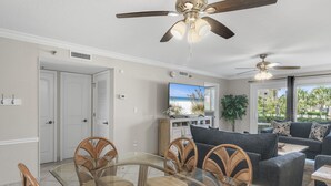 Uma sala de estar espaçosa com mesa de jantar de vidro, cadeiras de vime, um sofá escuro e ventiladores de teto.