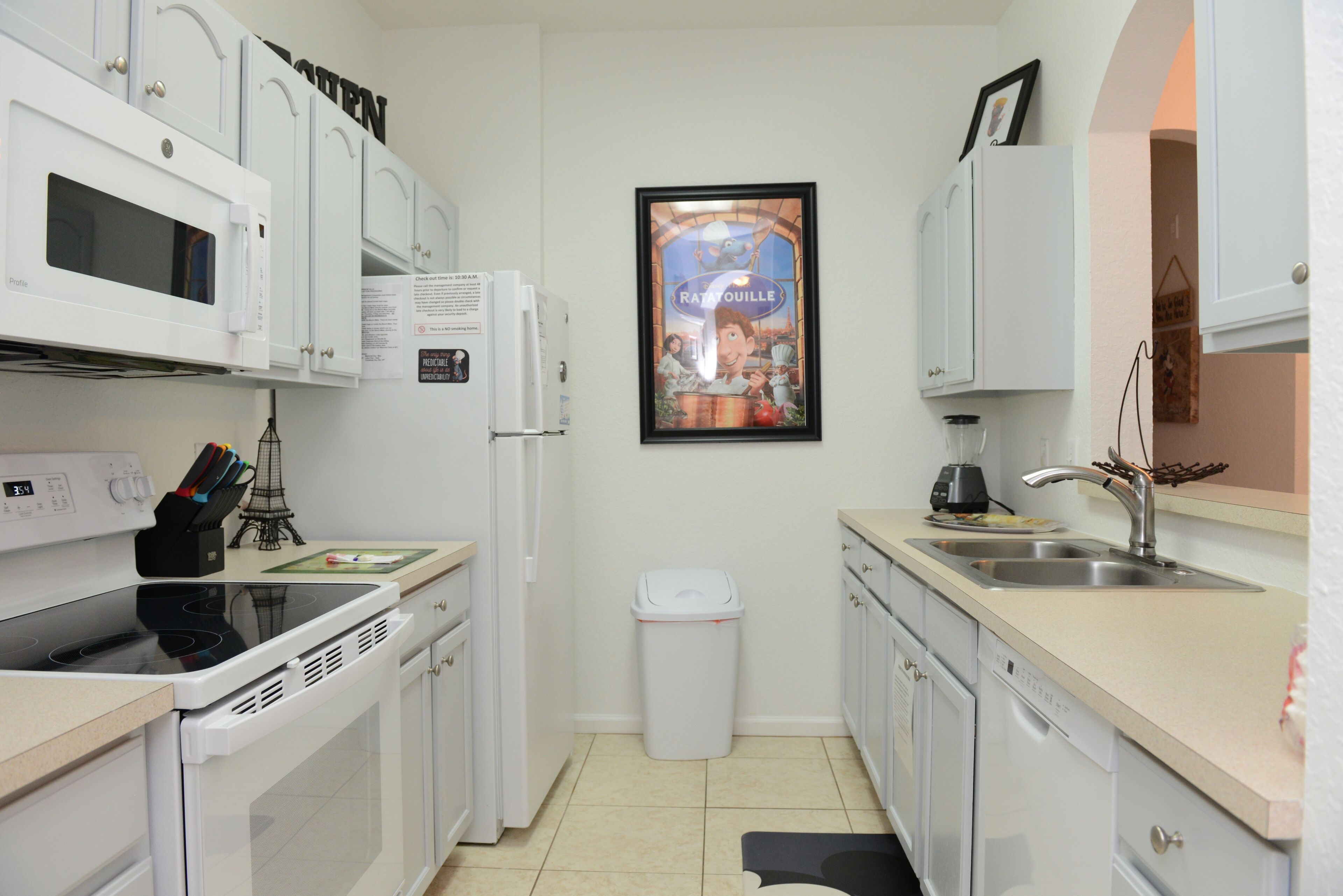 A compact kitchen with white appliances, a trash bin, and a framed poster on the wall.