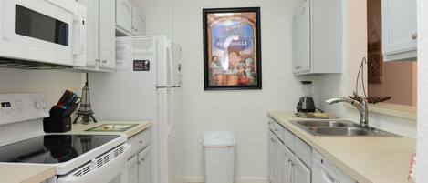 A compact kitchen with white appliances, a trash bin, and a framed poster on the wall.