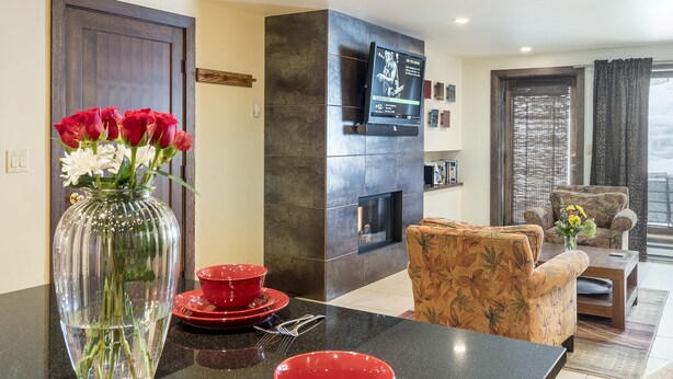 A living room with a fireplace, a TV mounted on the wall, and a dining area with red plates and a vase of flowers. - Walk to Thunderhead Lift! Hot Tub/ Pool! Quiet Building! (Steamboat Springs)