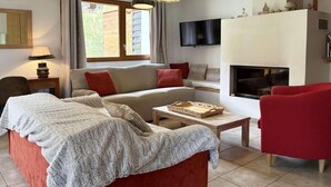 Een woonkamer met een rode bank, een beige bank, een rode fauteuil, een houten salontafel en een open haard.