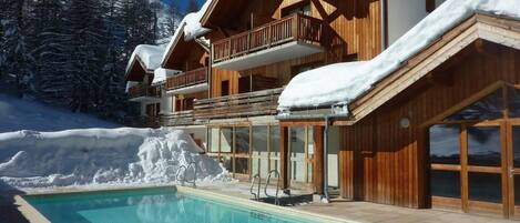 Un chalet en bois avec une piscine, entouré d’arbres couverts de neige et sous un ciel dégagé.