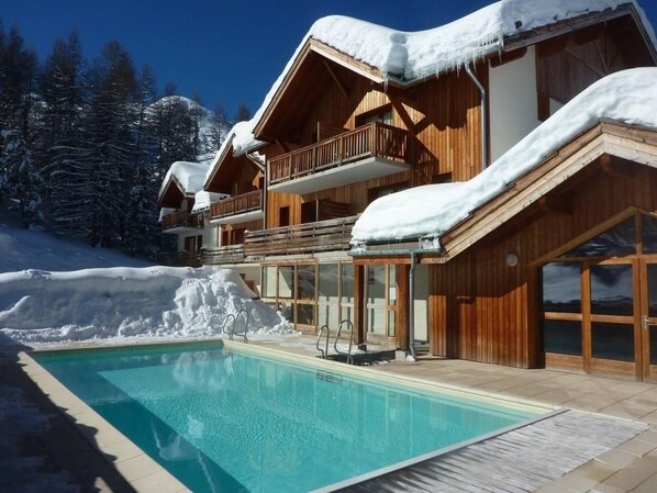 Una cabaña de madera con piscina, rodeada de árboles nevados y un cielo despejado.