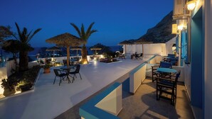 An outdoor terrace with tables and chairs, palm trees, and a view of the sea. - Ancient Thera Apartments (Santorini)