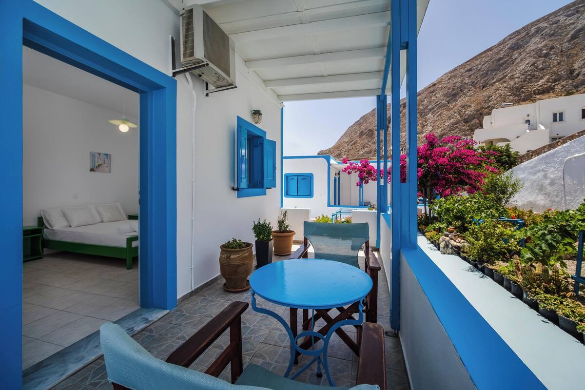 A balcony with a blue table and chairs, potted plants, and a view of a hillside.