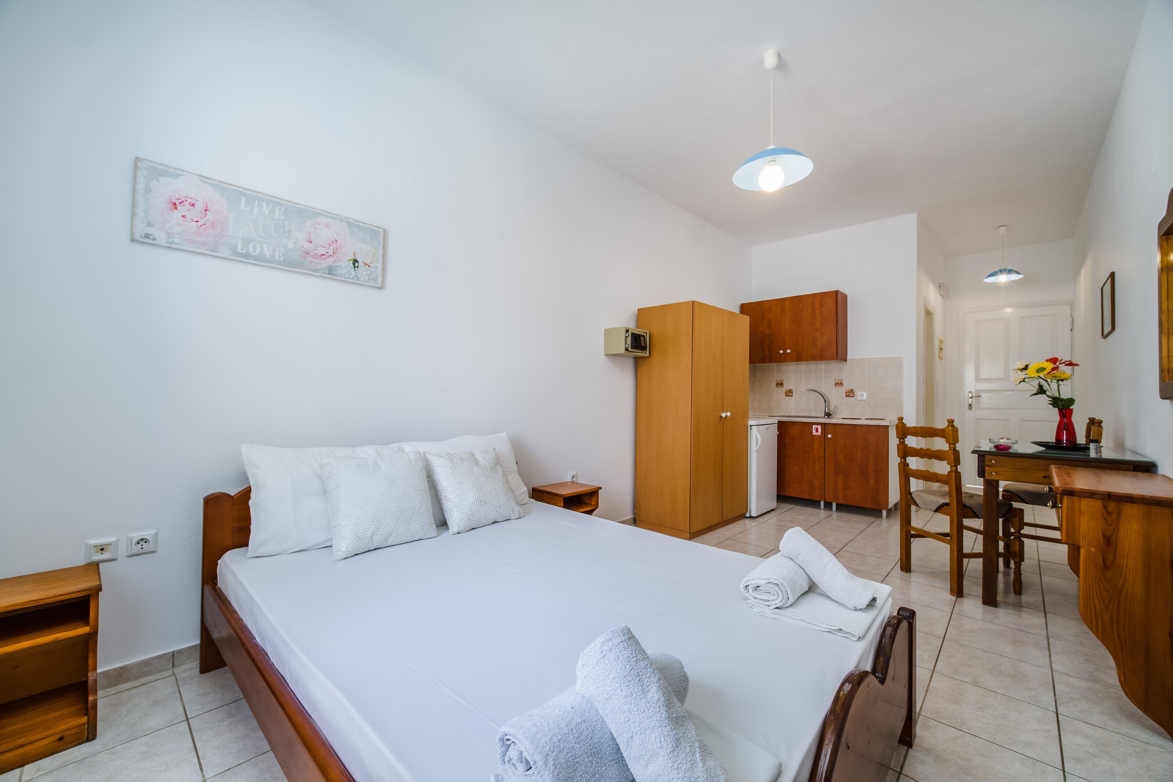 Un dormitorio con una cama grande, una mesita de noche de madera, una pequeña mesa y una zona de cocina al fondo.