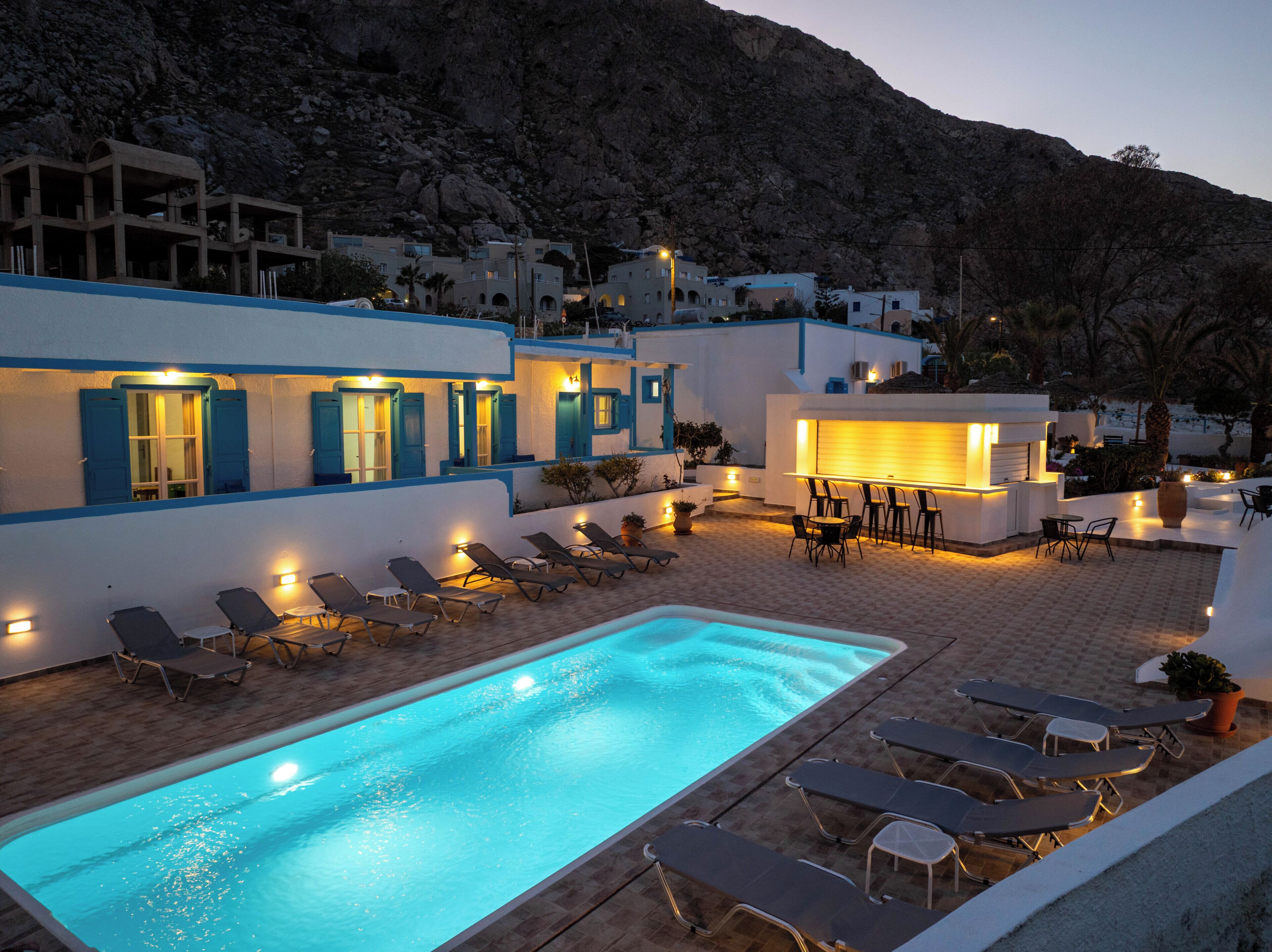 A poolside area with lounge chairs and a pool, surrounded by buildings with blue accents and a mountainous backdrop.