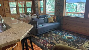 A cozy cabin interior with a stone wall, wooden furniture, a blue sofa, a patterned rug, and a large window with a view of greenery. - Cozy and comfortable wooded cabin (Alta)