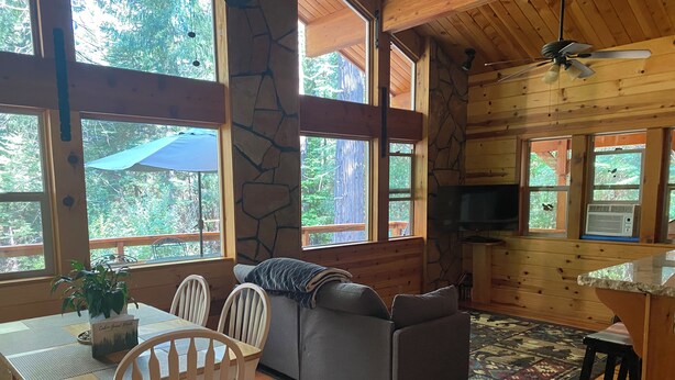 A cozy cabin interior with a dining area, a gray sofa, wooden floors, stone accents, and large windows offering a view of the outdoors. - Cozy and comfortable wooded cabin (Alta)