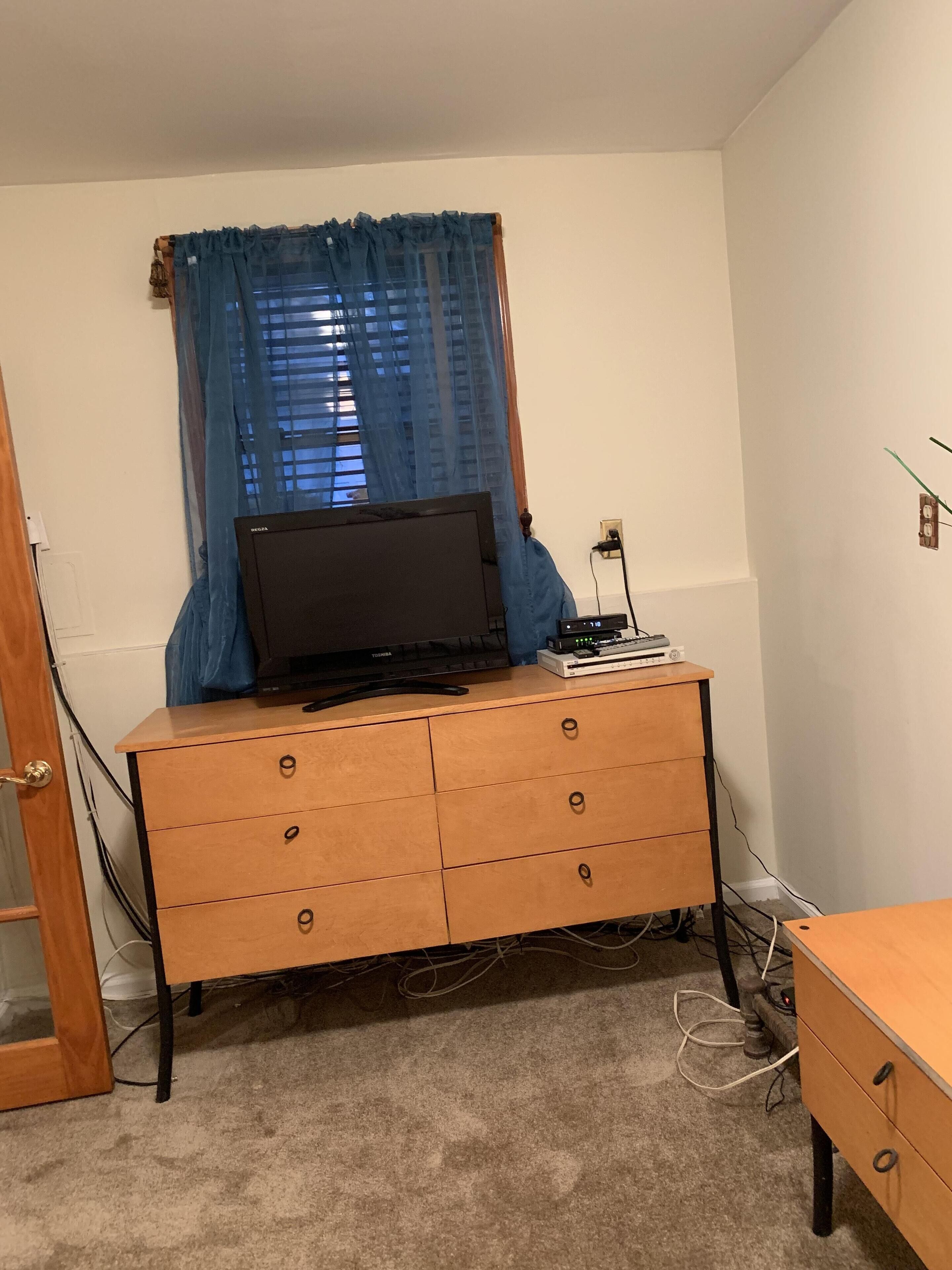 A room with a wooden dresser, a television on top, and a window with blue curtains.