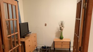 A room with a wooden door, a small wooden desk, a television on a stand, a vase with dried flowers, and a power strip on the floor.