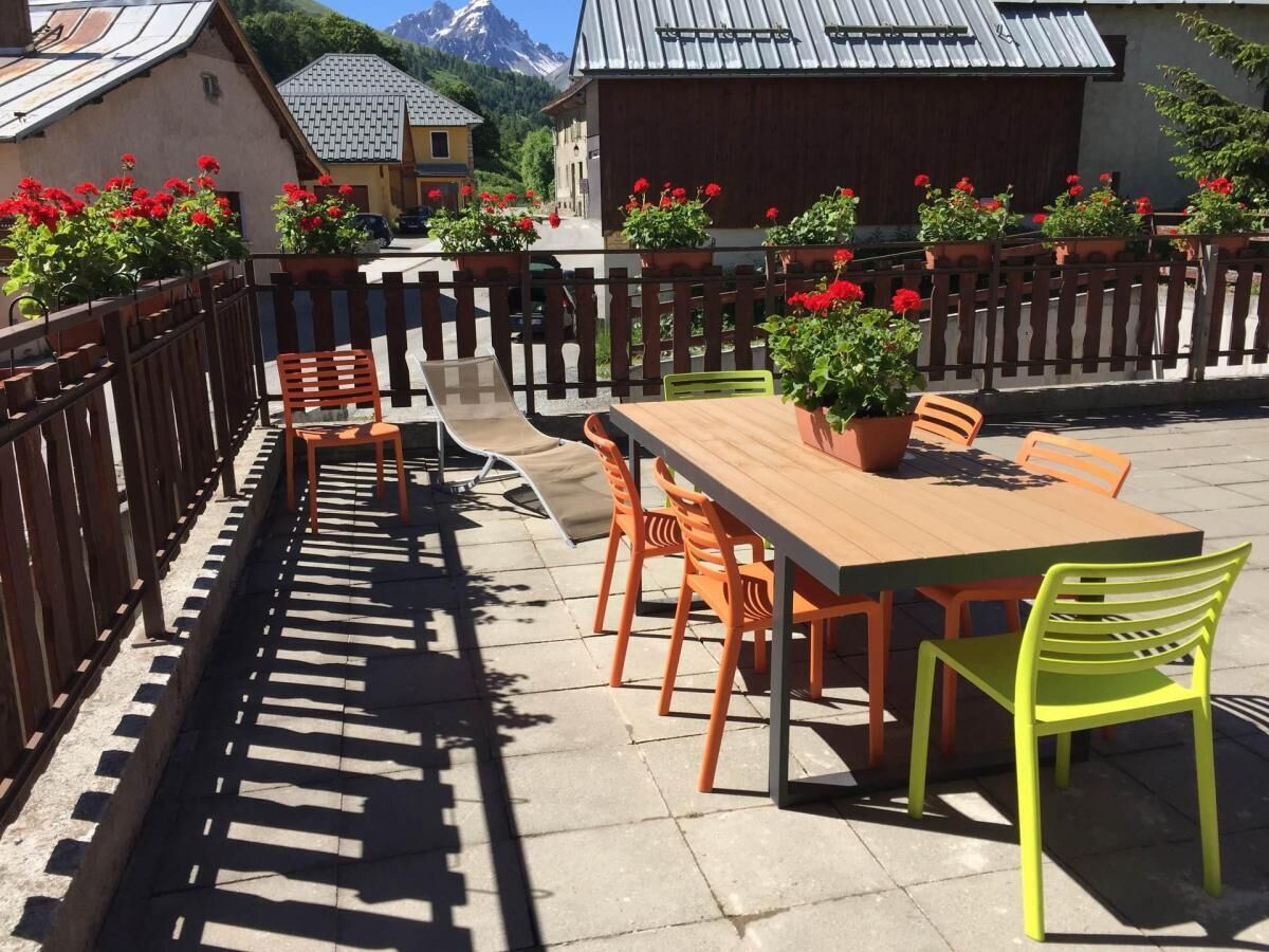 Een terras buiten, met een lange houten tafel en stoelen, omringd door een houten hek en plantenbakken.