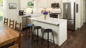 Uma cozinha moderna com armários brancos, eletrodomésticos de aço inoxidável, uma ilha central com banquetas e um espaço de jantar com mesa e cadeiras de madeira.